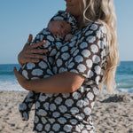 Woman holding a baby wrapped in a brown wrap with white shell patterns on a beach.