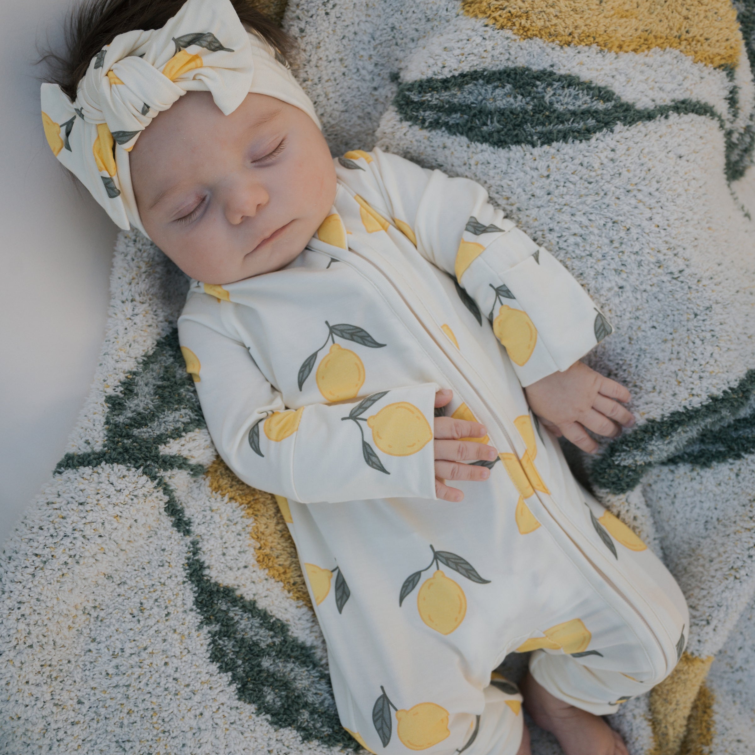 Baby wearing Lemon Bamboo Zip Pajamas, featuring a lemon print and matching headband, resting on a cozy blanket.
